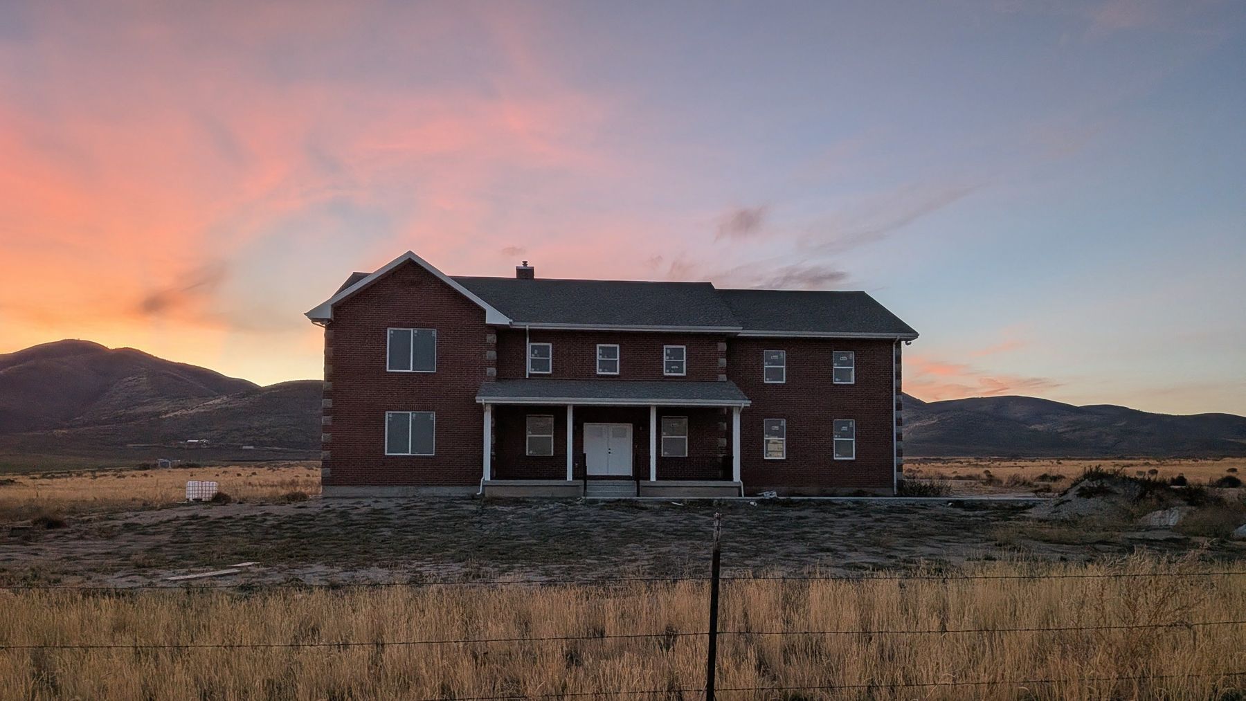 Two-story brick home with a covered front porch under a pink and blue sunset sky, hills behind.