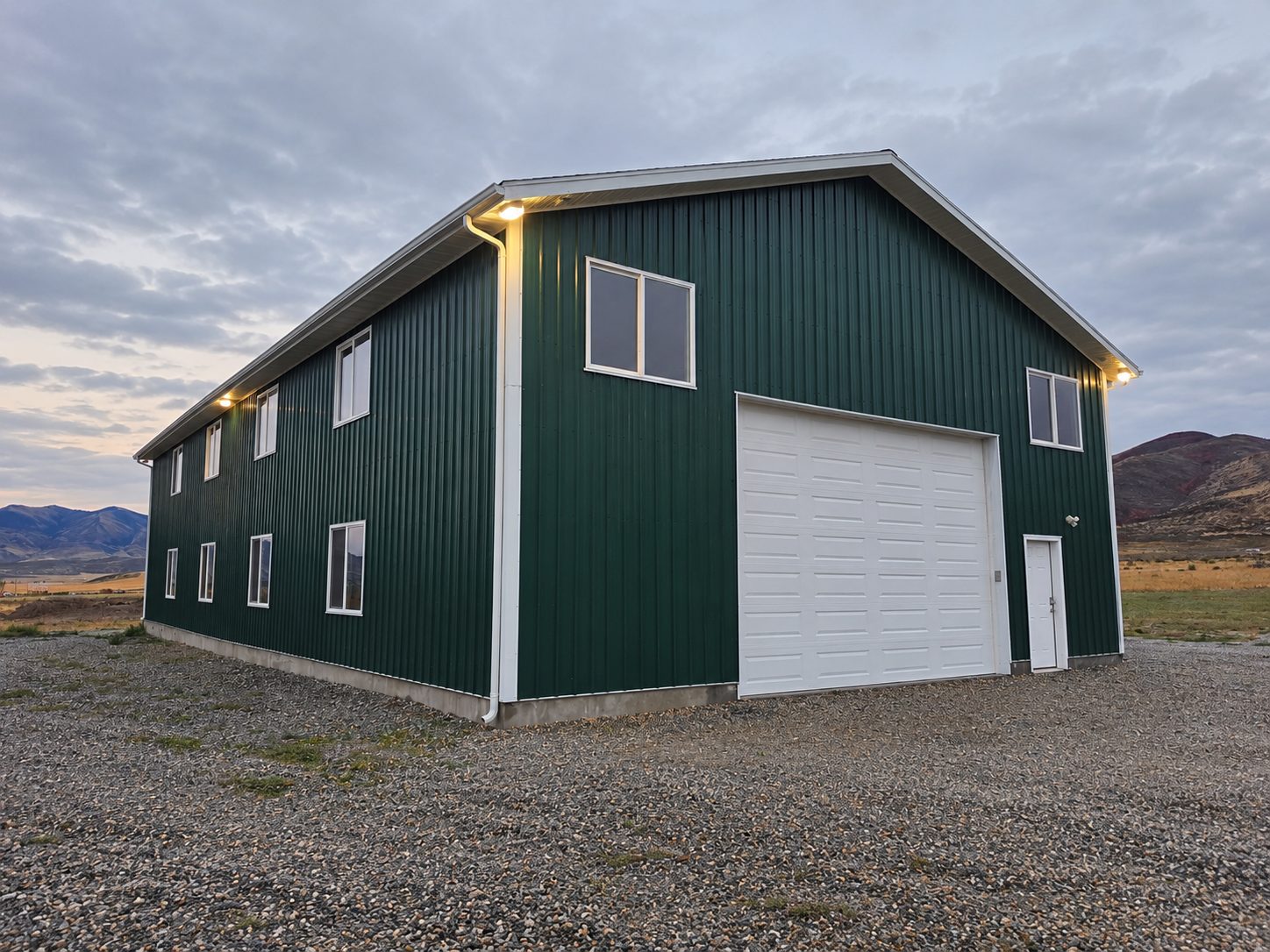 Green two-story metal shop building with a large roll-up door and gravel apron.
